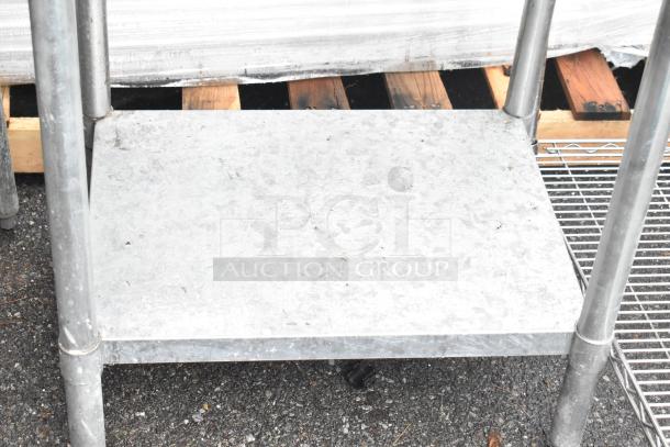 Stainless steel table with metal under shelf, includes wire shelving unit. Visible signs of wear; sturdy construction.