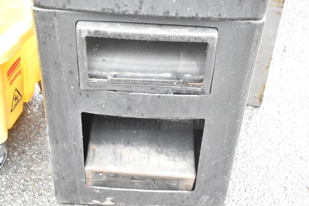 Black poly trash can with two compartments, showing signs of wear. No visible labels or markings.