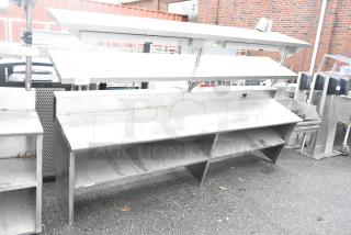 Stainless steel commercial table with two food strip warmers, under shelves, and over shelves, showing visible wear.