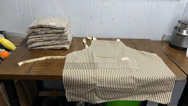 Striped aprons on table, brand new condition, featuring a front pocket and beige ties. Bulk set ready for sale or use.