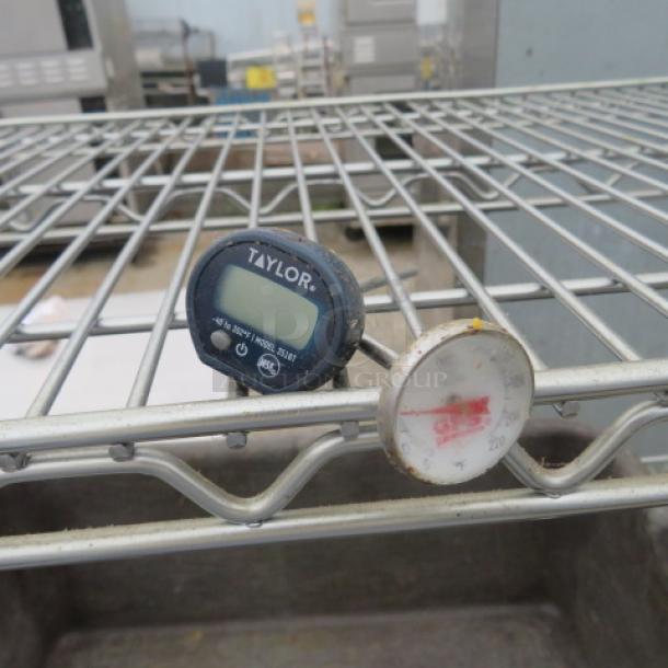 Two assorted thermometers on a metal shelf: a digital Taylor thermometer and an analog one, both in used condition.
