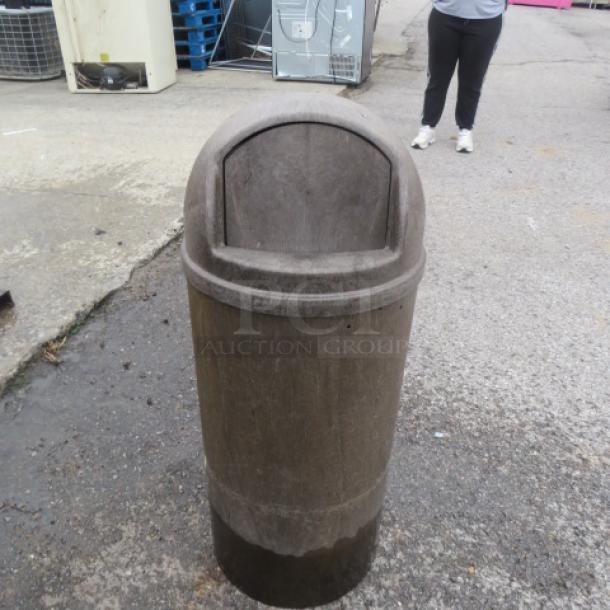 Heavy-duty round trash can with flip lid, used condition, outdoor setting. Durable construction for high-traffic areas.