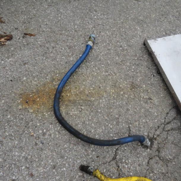 Blue gas line with metal fittings on each end, resting on concrete. Slight discoloration on surface. Used condition.