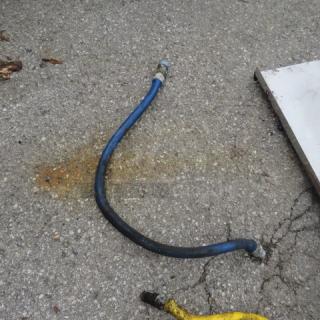Blue gas line with metal fittings on each end, resting on concrete. Slight discoloration on surface. Used condition.