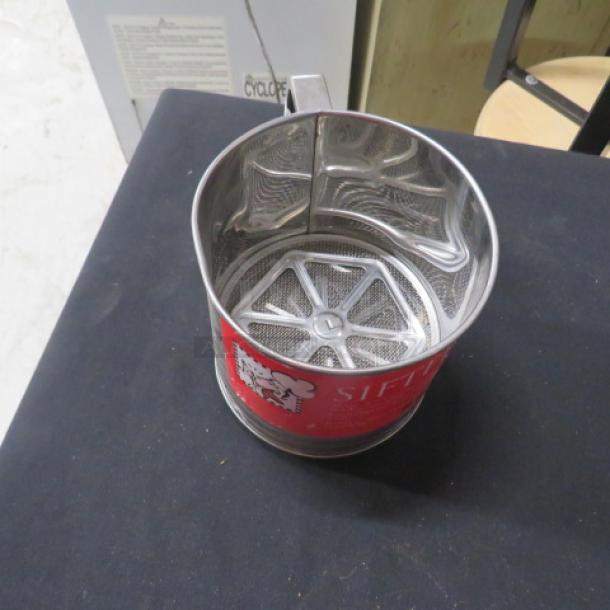 Stainless steel sifter with crank handle, red label, mesh base. Mint condition, labeled "NEW." Perfect for baking.