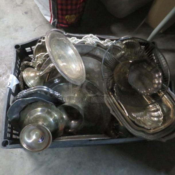 Assorted silver-plated serveware in a plastic container, featuring trays, bowls, and platters. Various styles and moderate tarnish.