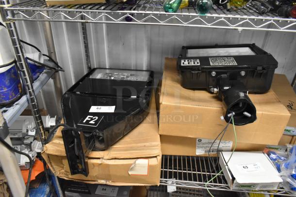 Four black flood lights on shelves with tags. Includes labels "F2" and "62 LED." Appears unused, in excellent condition.