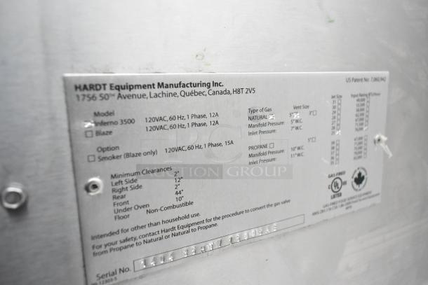 Hardt Inferno 3500 stainless steel rotisserie oven, commercial-grade with skewers and metal legs on casters. Serial label visible.