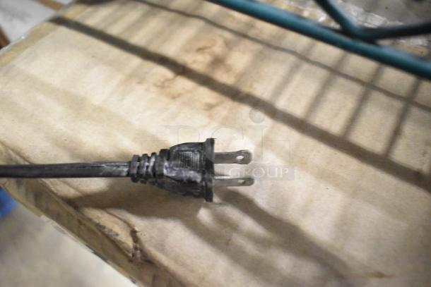 A black two-prong power plug on a cardboard surface. No air fryer oven visible.