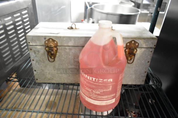 Metal toolbox with brass latches and red jug of sanitizer on a shelf. Items in used condition, toolbox has a small label.