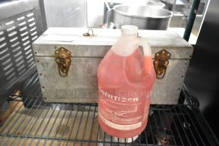 Metal toolbox with brass latches and red jug of sanitizer on a shelf. Items in used condition, toolbox has a small label.