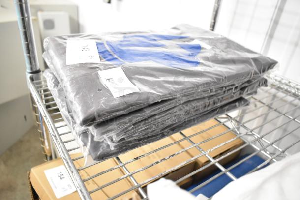 6 brand new Uncommon Chef slate gray chef shirts, scratch and dent condition, in packaging with tags on metal shelf.