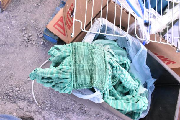 Black Poly Slim Jim trash can filled with assorted green fabric and cardboard boxes. Some metal shelving parts visible. Item is used.