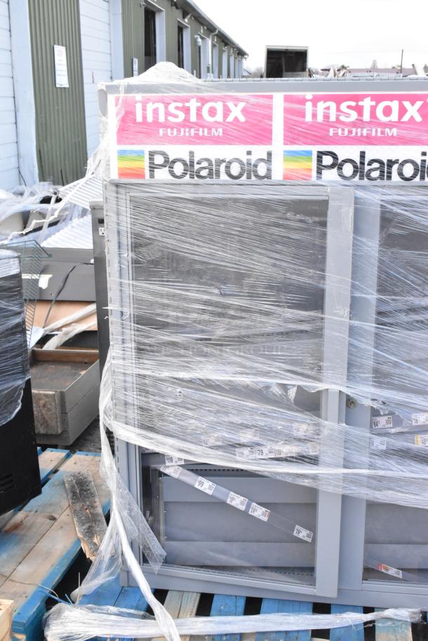 Gray metal cabinet wrapped in plastic, featuring Fujifilm Instax and Polaroid branding, visible pricing labels.
