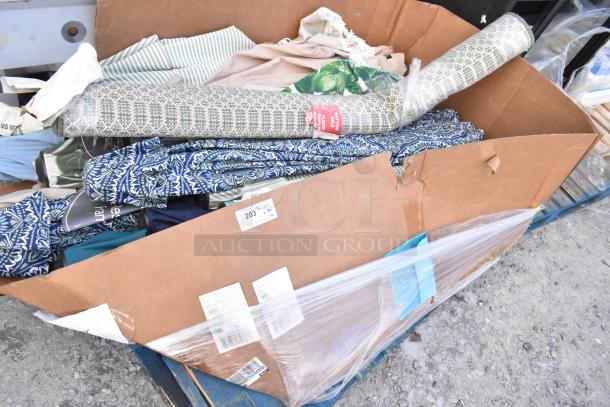Cardboard box with various items including fabric rolls and patio umbrella components, lightly used. Barcodes and labels visible.