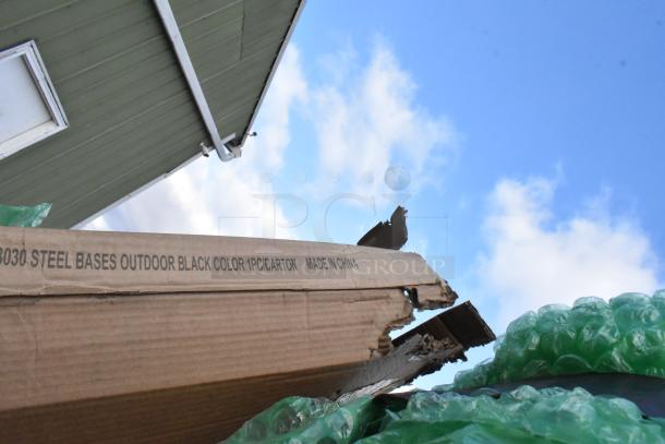 Cardboard box labeled "Steel Bases Outdoor, Black, Made in China," with visible damage. Green bubble wrap visible.