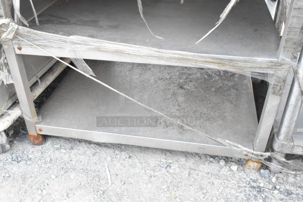 Stainless steel equipment stand with two shelves, wrapped in plastic. Rust visible on base. Suitable for heavy-duty use.