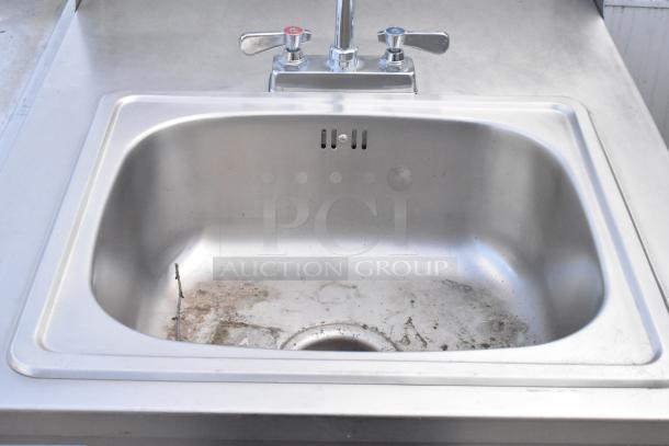 QualServ stainless steel commercial portable sink with dual faucets, on casters. Good condition, some dirt visible in basin.
