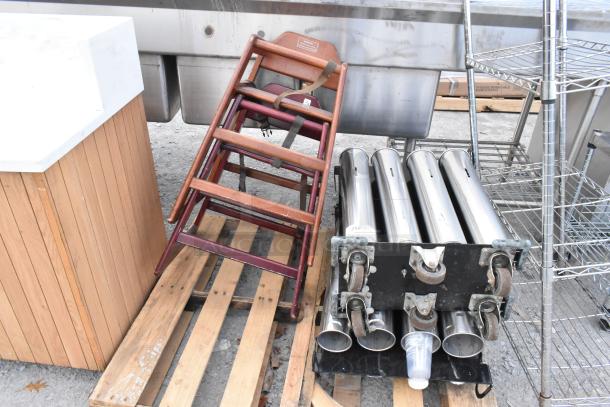 Wooden high chair with safety straps and metal cup dispenser on a pallet, showing signs of use.