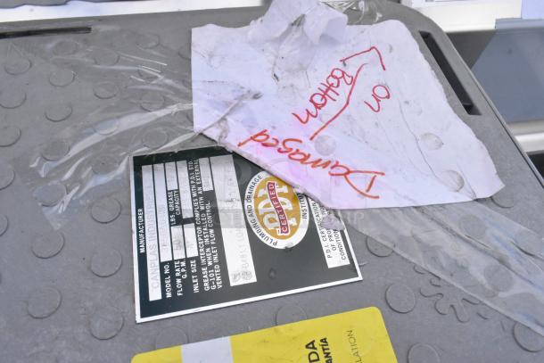 Grey grease trap lid with label showing model G99, 50 lbs capacity, from Canplas Industries Ltd. Damaged tag on top.