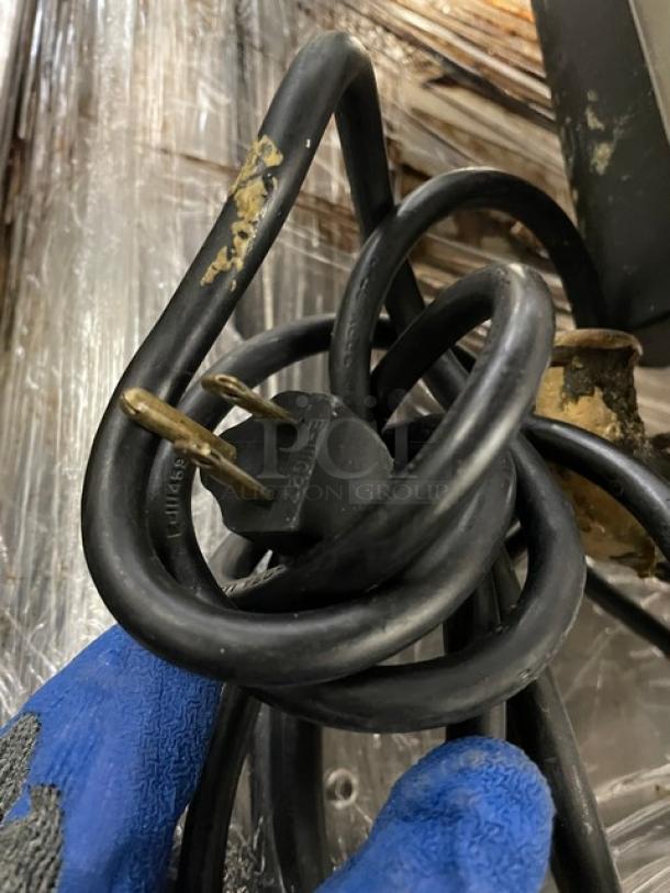A coiled black electrical cord with a standard plug on a worn background, next to a blue-gloved hand.