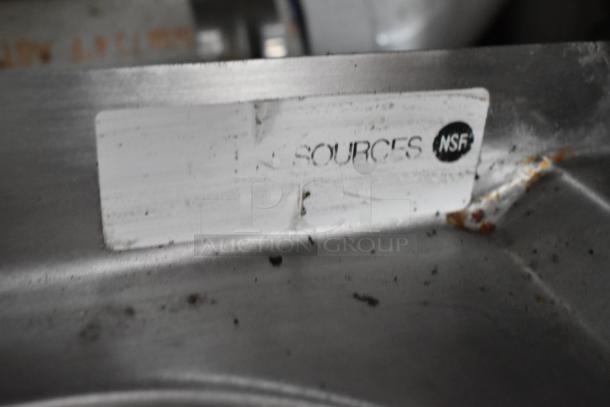 Close-up of a metal surface with a label marked "SOURCES" and "NSF," indicating certification. Small scuffs and dirt visible.
