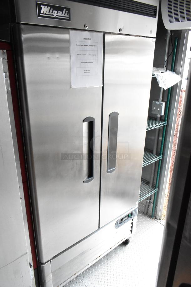 Stainless steel Migali refrigerator inside fully equipped mobile kitchen trailer, featuring attached inventory list and shelving.
