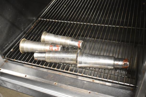 Four cylindrical metal parts inside an industrial oven, resting on a metal rack.
