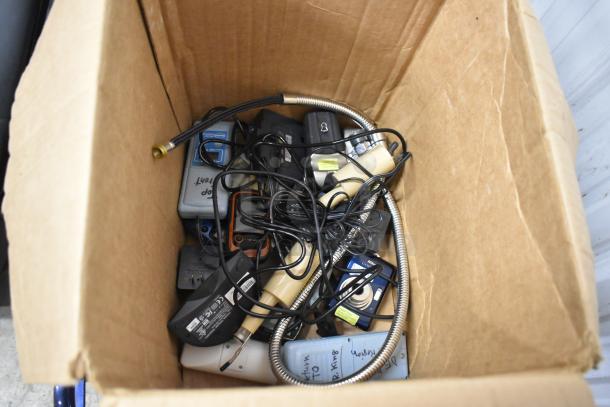Cardboard box containing assorted electronics, including digital cameras, Garmin GPS, and Hach colorimeters, numerous cables and parts, mixed condition.