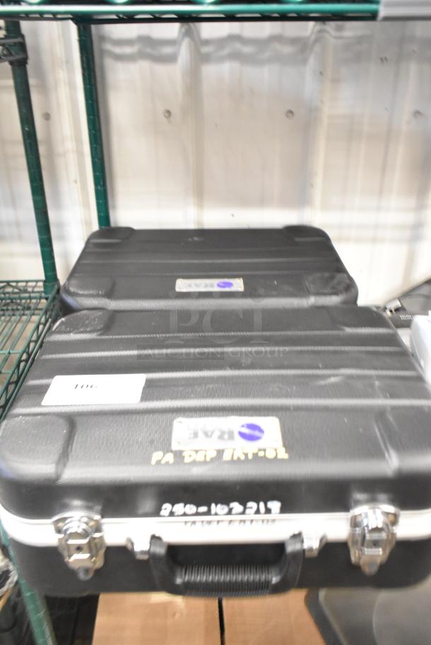 Two black hard cases on a shelf, labeled with "RAE" and "PA DEP EAT-02." Secure metal clasps visible, sturdy condition.