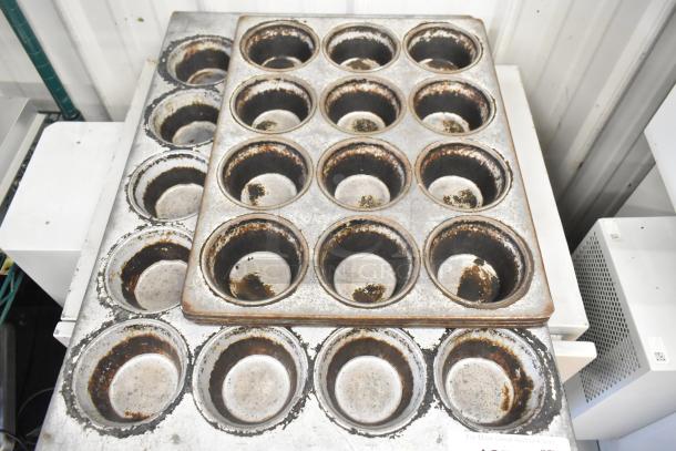 Seven metal muffin baking pans, including 12-cup and 20-cup pans, showing signs of wear.