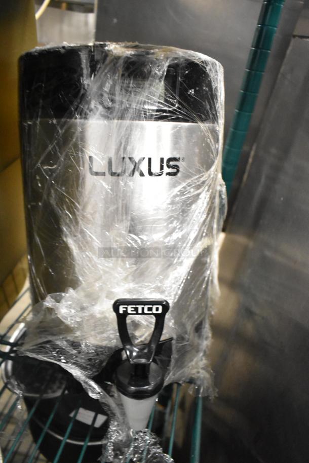 Fetco Luxus beverage dispenser, wrapped in plastic, stainless steel finish, visible branding. Includes tap for dispensing.