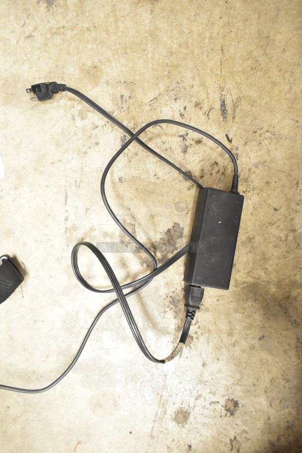 Image of a power adapter on a concrete surface, possibly for an HP All In One Desktop, showing wear. Cables and plug visible.