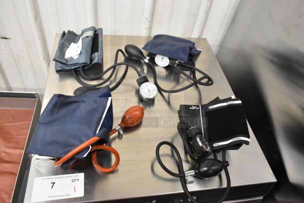 Four assorted blood pressure cuffs with pumps and gauges on a table; mixed colors and styles, in used condition.