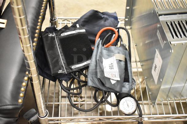 Four assorted blood pressure cuffs on a metal shelf, varying in size and design, featuring Velcro straps and pressure gauges.