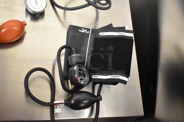 Four assorted blood pressure cuffs on a metal surface, featuring black cuffs, hoses, and gauges. Text on cuffs indicates size range.