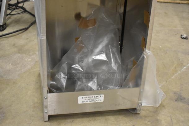 Metal box with shipping brace indicated on label, partially wrapped in plastic, resting on concrete floor.