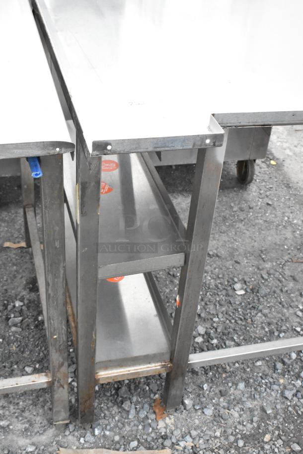 Stainless steel table with two shelves, visible bolts, minor wear, on gravel ground.