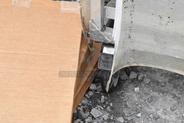 Metal commercial pan transport rack corner on casters with brake, covered by mesh fabric, on gravel surface.