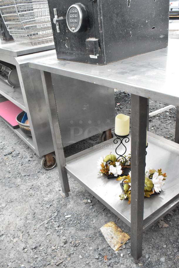 Stainless steel table with two shelves, showing light scuffs. Features a sturdy metal frame and casters for mobility.