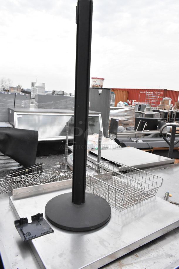 Stainless steel table with metal basket and black base, in used condition, outdoor setting. Ideal for commercial use.