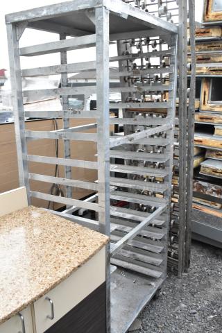 Metal commercial pan transport rack with multiple shelves on heavy-duty casters, showing signs of wear. Ideal for bakery use.