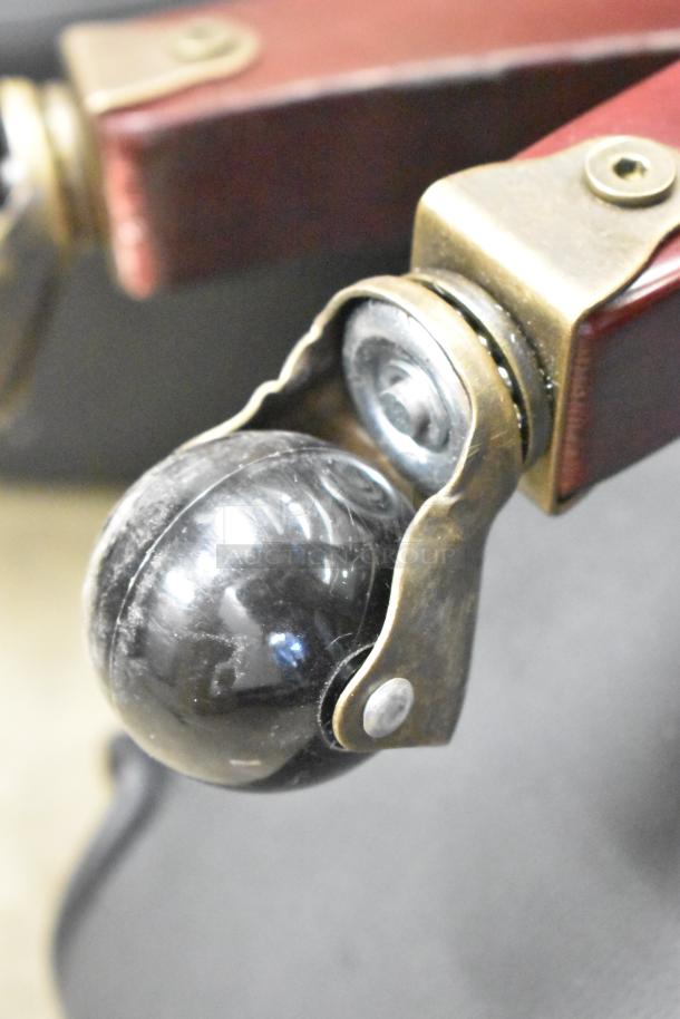 Chair caster wheel close-up, featuring a black spherical wheel in a brass housing attached to a red armrest. Good condition.