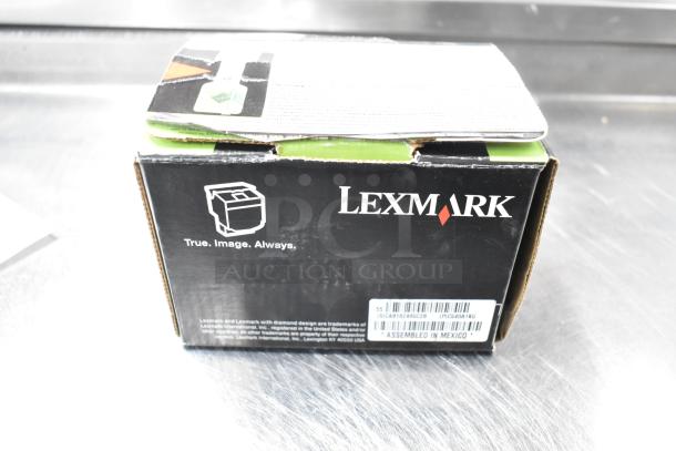 Lexmark printer cartridge box with branding, barcode, and "Assembled in Mexico" text, part of a lot including notebooks and paper.