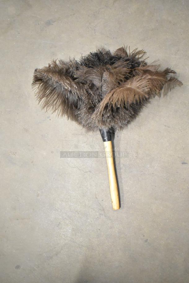Feather duster with wood handle and dark brown feathers, shown on concrete floor.
