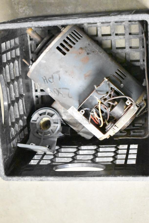 A black crate contains various metal components including parts of a motor and wiring, tagged with handwritten markings, possibly used.
