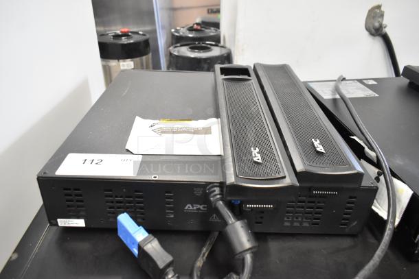 Two APC uninterruptible power supplies, black, with visible cables and labels. Includes notice and identification tags.