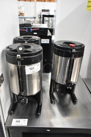 Three Fetco metal countertop beverage dispensers, stainless steel and black finish, with spouts. Visible tags indicate quantities and lot numbers.