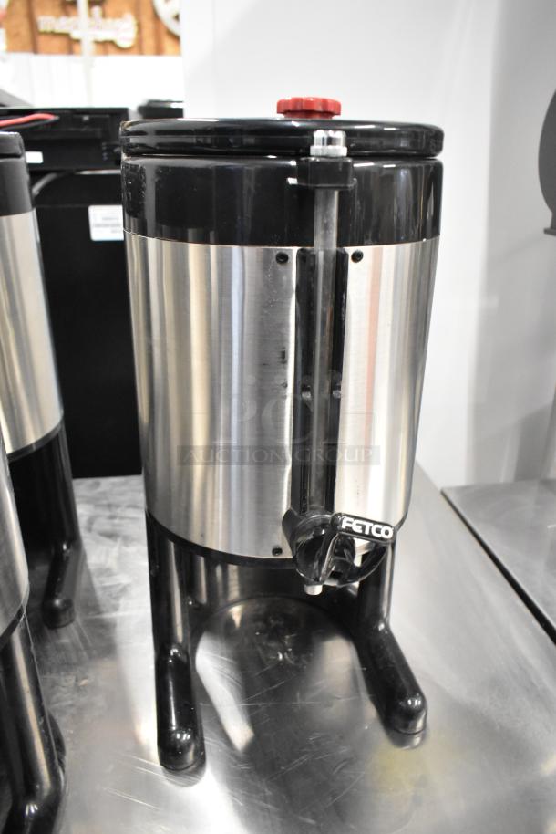 Fetco metal countertop beverage holder dispenser, stainless steel with black accents, featuring a dispensing spout and red top knob.