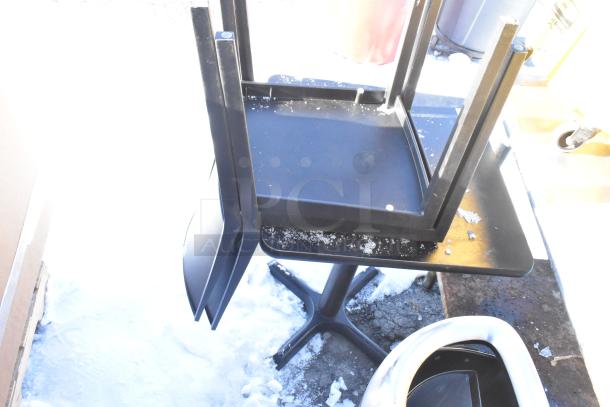 Black table with pedestal base and two stackable chairs, visibly worn, outdoor setting.